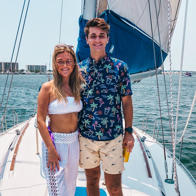 Smiling young couple standing on a sailboat deck during a sunny cruise, wearing summer vacation outfits with the ocean in the background.