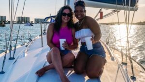 Two women sitting on the deck of a private sailing charter, smiling and holding drinks, dressed in casual summer outfits