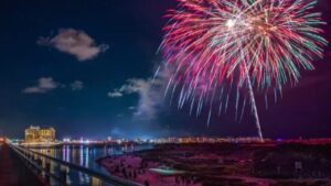 Fireworks bursting over a waterfront at night, with a lit pier and people below.